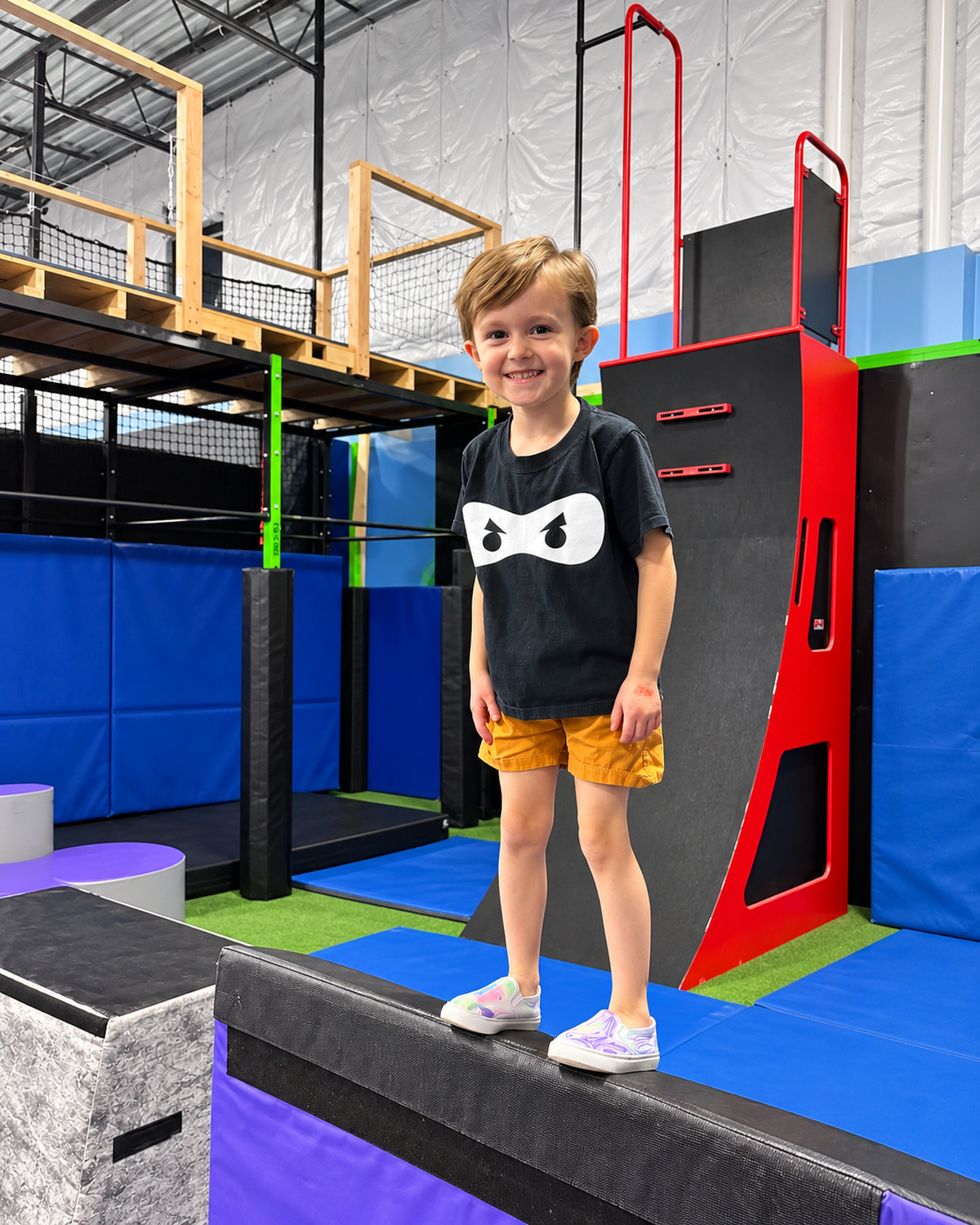 Smiling boy in a ninja shirt standing on a beam in the ninja course