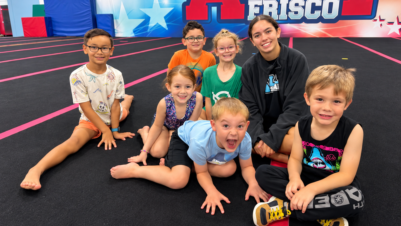 A group of smiling campers sitting with a coach on the cheer floor at All 4 Frisco