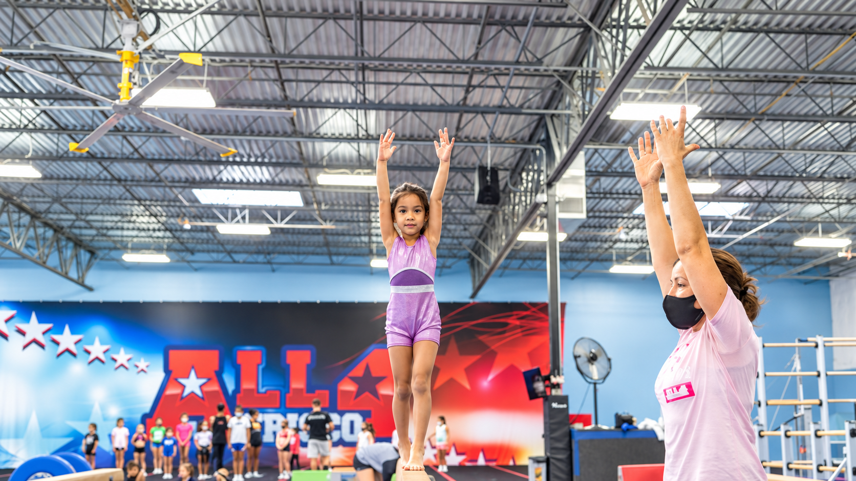 A young gymnast balancing on the beam with arms raised while a coach mirrors her pose