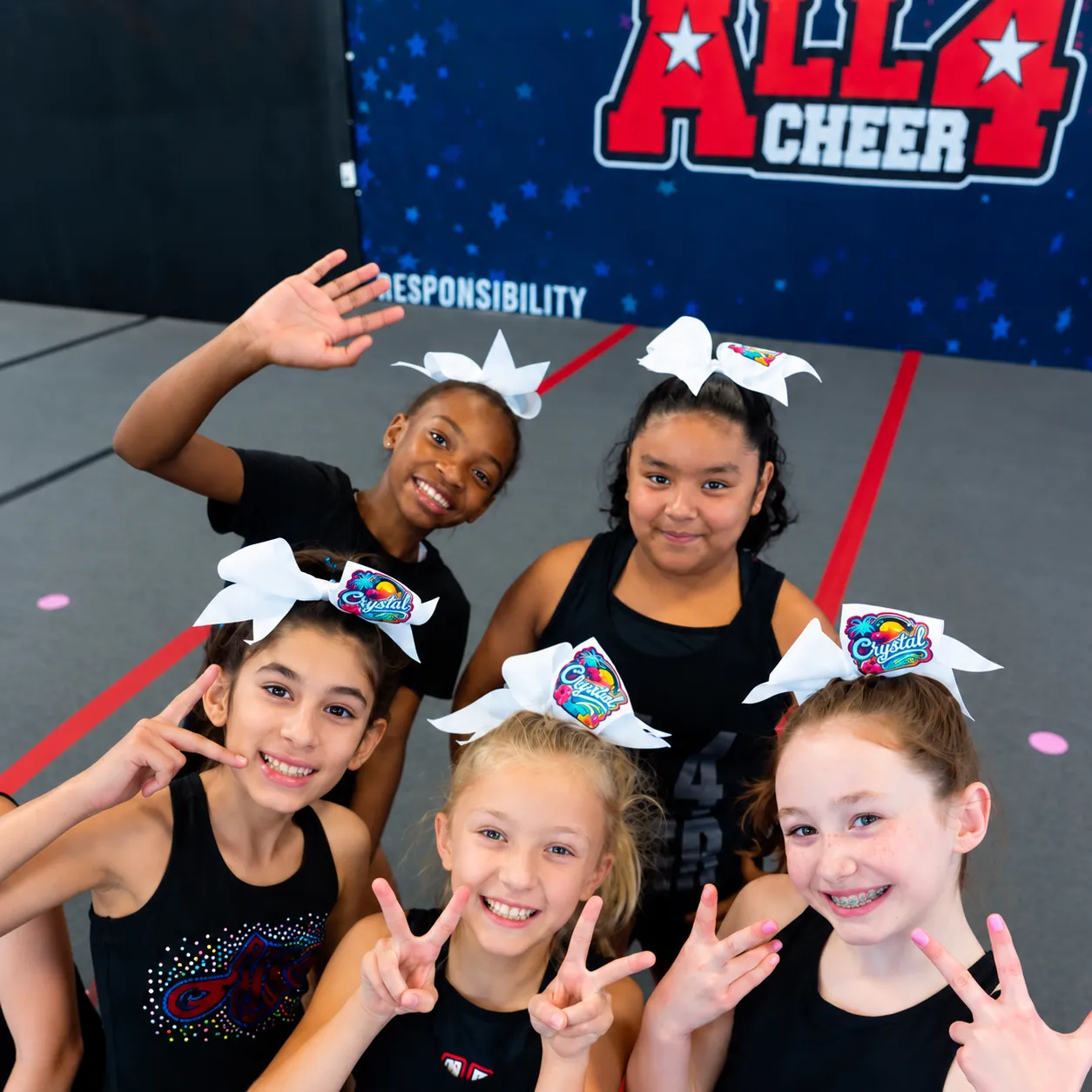 Five cheerleaders in white bows waving and making peace signs in front of the All 4 Cheer backdrop
